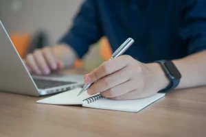 Mand skriver noter og sidder med sin laptop, hvor han vil skrive et nyt blogindlæg